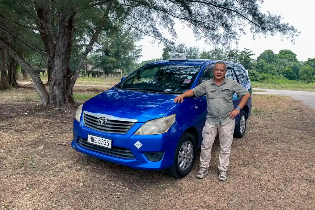 Driver standing beside a blue Toyota Innova taxi on a roadside in Malaysia