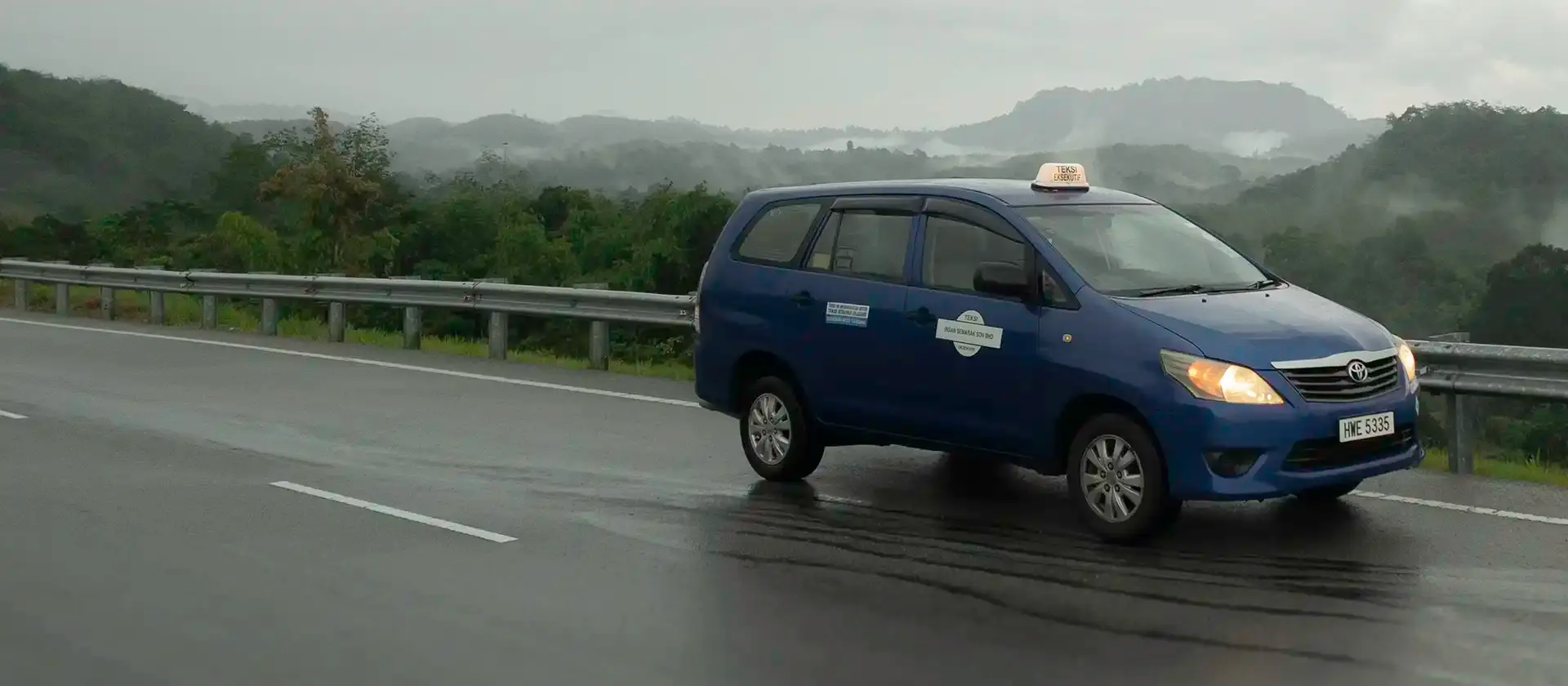 Blue Toyota Innova taxi on a road with forested hills in the background.