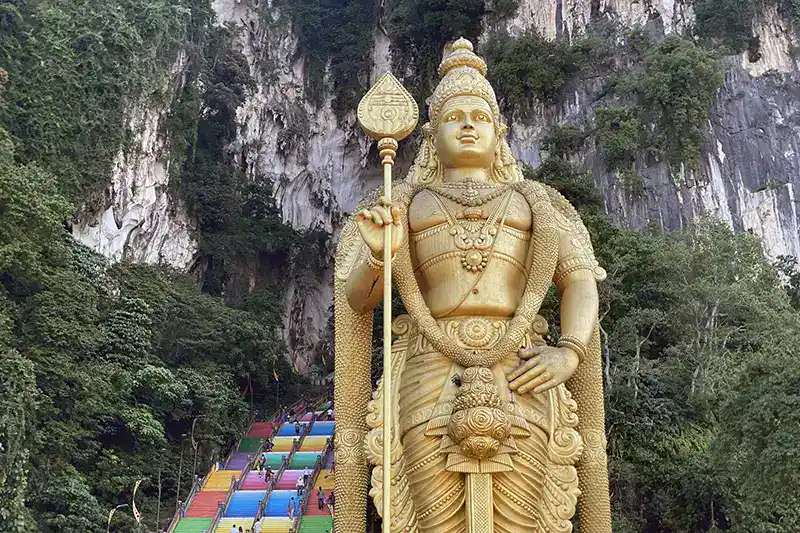 The golden Lord Murugan statue at Batu Caves with the colourful staircase and limestone cliffs behind it.