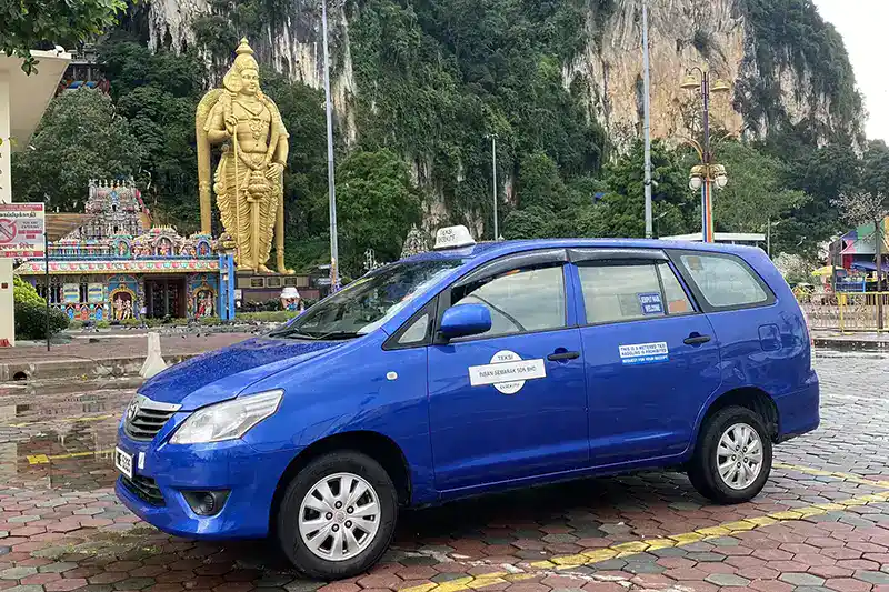 Blue taxi parked at Batu Caves with the Lord Murugan statue and temple in the background.