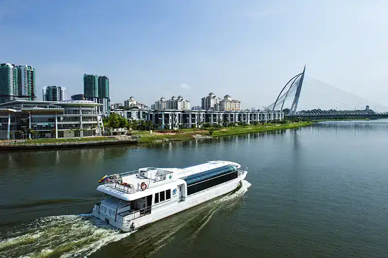 A sightseeing boat cruising along the lake in Putrajaya with modern buildings and a cable-stayed bridge in the background.