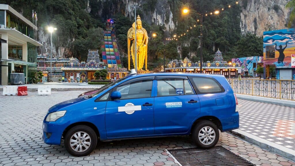 Blue Toyota Innova private hire transfer parked at Batu Caves temple with the Murugan statue and rainbow staircase behind