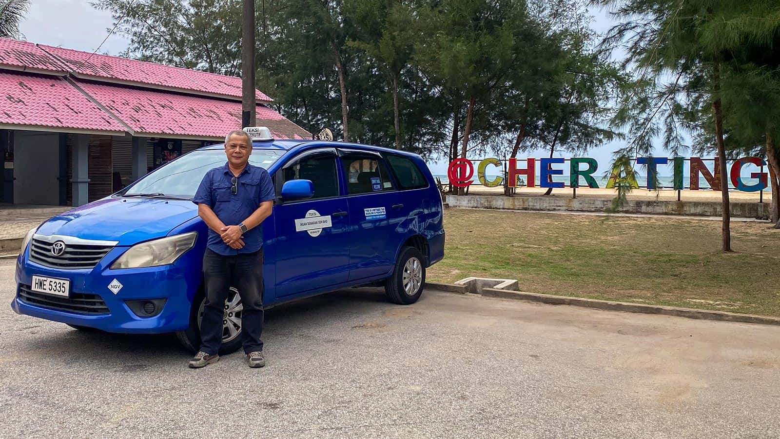 Anuar standing beside his blue Toyota Innova taxi at the Cherating beach signage after a private transfer from Kuala Lumpur